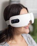 Woman wearing white electric eye massager with digital control panel, using heat, vibration, and air pressure therapy for relaxation and eye strain relief