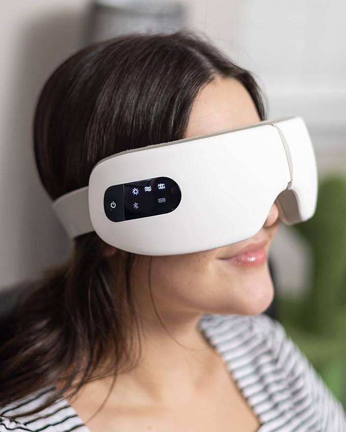 Woman wearing white electric eye massager with digital control panel, using heat, vibration, and air pressure therapy for relaxation and eye strain relief