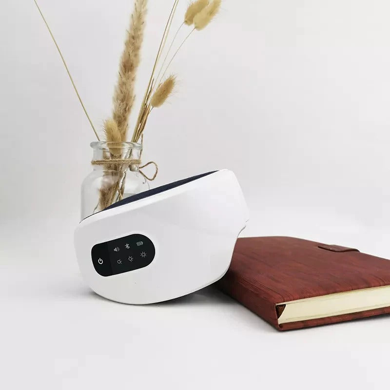 White rechargeable electric eye massager with LED control panel placed beside a notebook and vase, designed for heat, vibration, and air pressure therapy to relieve eye fatigue and improve sleep